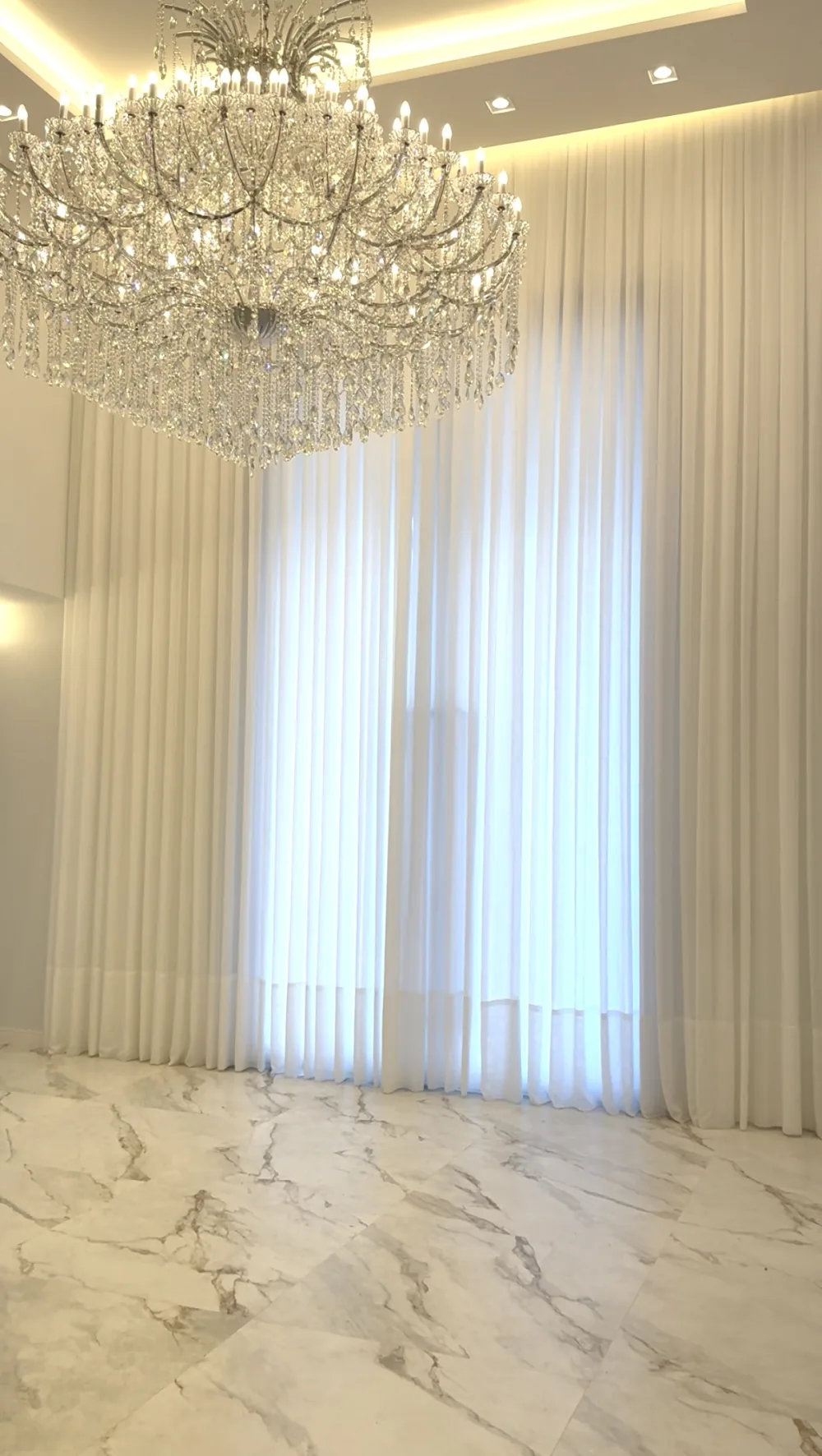 Sala de pé-direito duplo com voil branco do teto ao chão cobrindo a janela inteira, lustre de cristal e piso de mármore.
