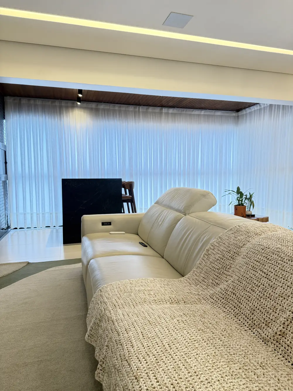 Sala de estar com sofá de couro claro e a janela panorâmica coberta por voil branco, que difunde a luz do fim de tarde.
