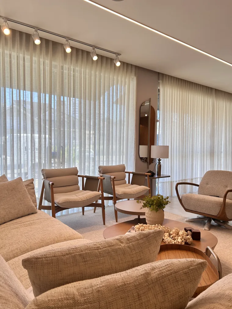 Sala do showroom com poltronas de madeira e assento estofado, espelho oval e abajur, diante de um voil claro de parede inteira.