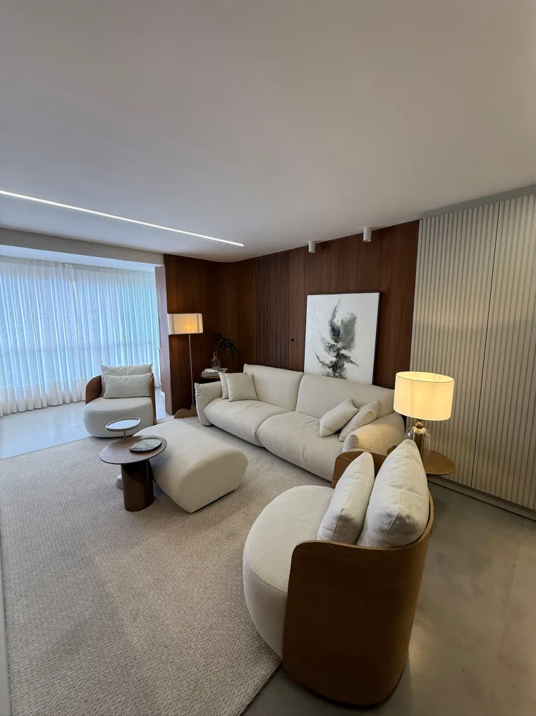 Ambiente com sofá claro comprido, poltrona de couro caramelo, painel de madeira e cortina de voil ao fundo.