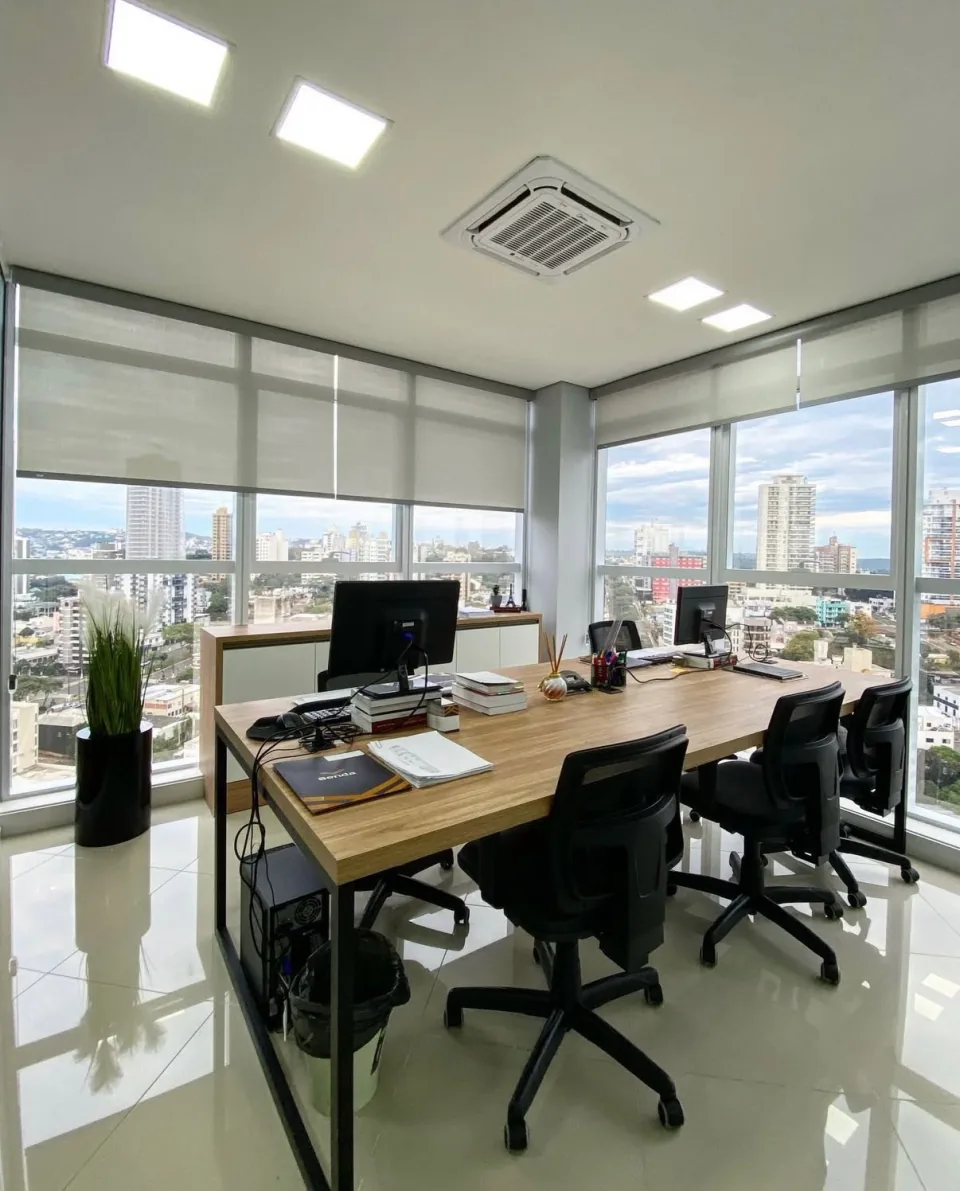 Sala de reunião envidraçada em andar alto, com persianas rolô claras recolhidas até a metade e a vista da cidade de Chapecó ao fundo.