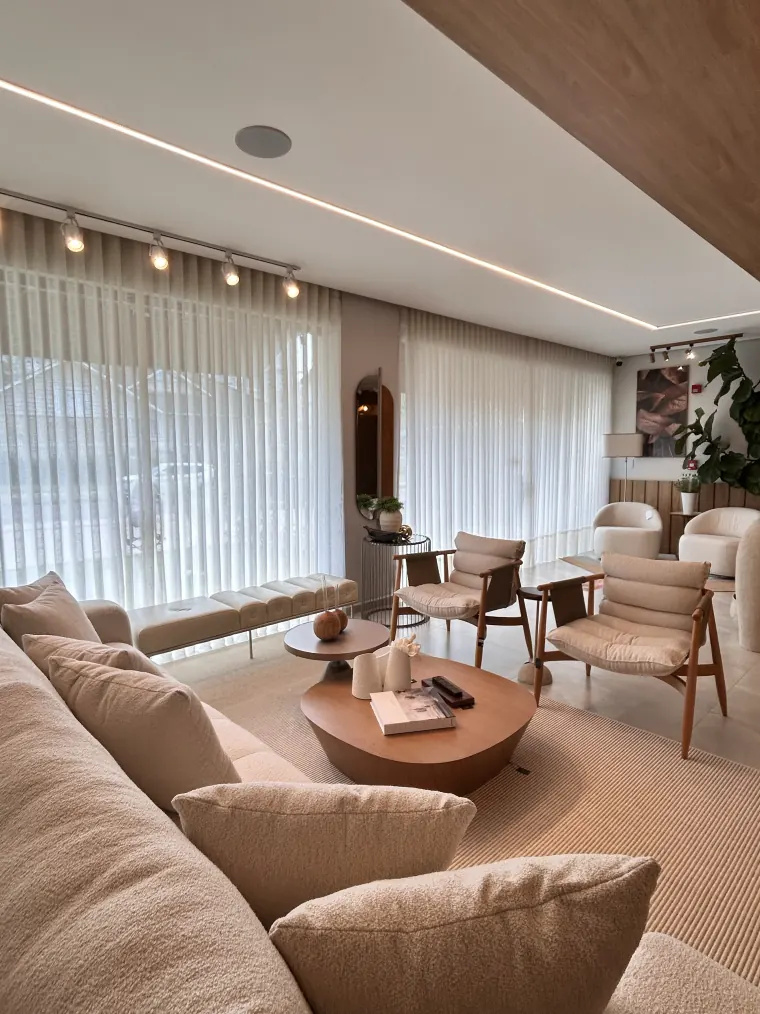 Vista ampla do showroom, com dois ambientes montados, sofás claros e cortinas de voil cobrindo a fachada de vidro.