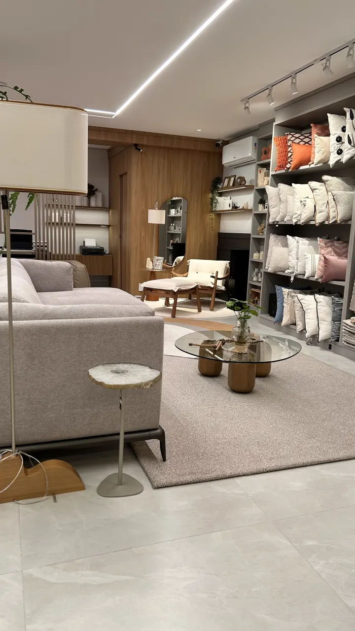Prateleiras do showroom cheias de almofadas de tecidos e cores diferentes, com tapete e sofá cinza à frente.