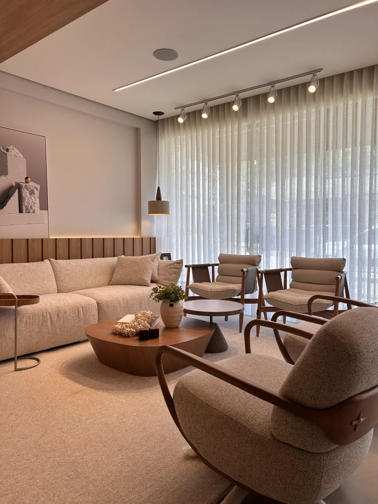 Ambiente montado no showroom da Tezzini, com sofá bege, poltronas de madeira e a parede inteira de voil claro atrás.